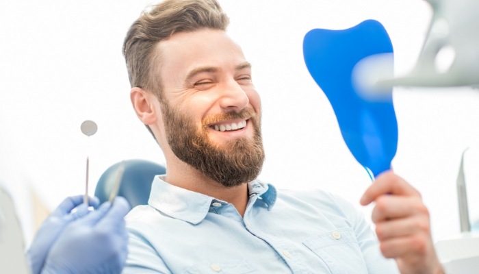 Young bearded man in dental chair seeing his smile in blue mirror