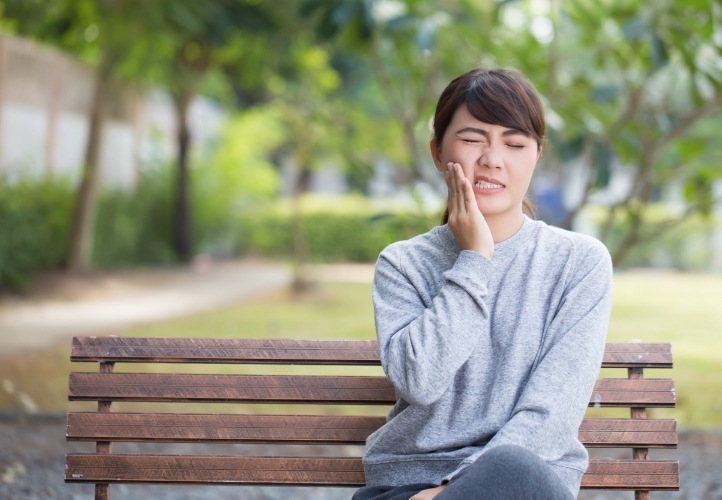 Woman who needs emergency dentist in Saint Peters sitting on bench and holding cheek in pain